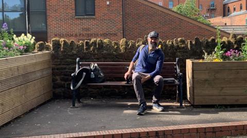 A man sitting on a wooden bench, smiling, looking at the camera and wearing a blue top, trousers, trainers and a cap. He has two large flower planters beside him.