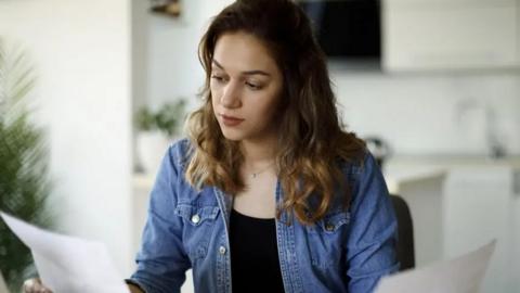 A photograph of a woman with brown hair of medium length looking at a piece of paper. 