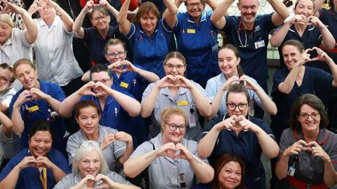 People dressed in NHS nurse outfits making heart gestures with their fingers at the camera. Most of them are female and dressed in uniforms ranging from grey to deep blue