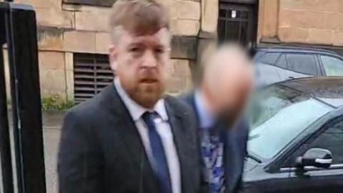 Kevin Montgomery - a man with ginger hair and a beard, arriving at court. He is dressed in a suit with a blue tie. Another man, whose face has been blurred, is walking beside him. 