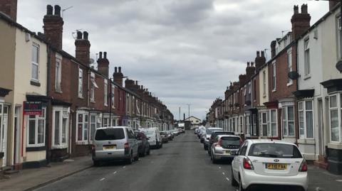 A street of terraced houses with bow windows on the ground floor and cars parked on both sides of the road