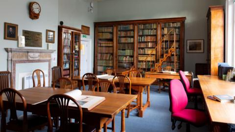 A reading room. There is a large brown book shelf with copious amounts of books on it.
There are also desks with chairs in front of the book shelf.
The floor has blue carpet and the walls are a light blue colour.