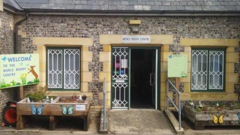 External view of the Merle Boddy Centre in Swaffham. The flint building has a ramp with grab rails leading to the green door. There are planters decorated with colourful butterflies outside. 