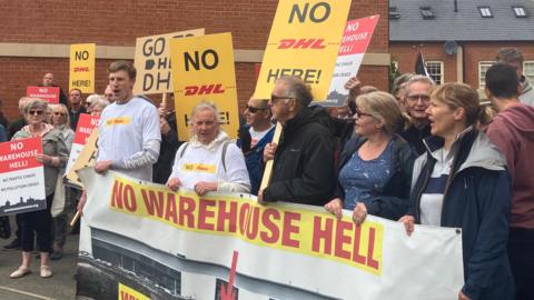 A line of men and women holding a banner saying "NO WAREHOUSE HELL" in red writing and placards with similar messages. Some appear to be shouting. Some have sweatshirts with a "NO DHL" slogan. They are standing in front of a brick building.