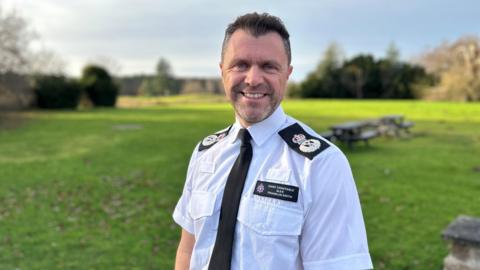 A man is looking at the camera and smiling. He is wearing a a white shirt and black tie and police labels and a name badge. 