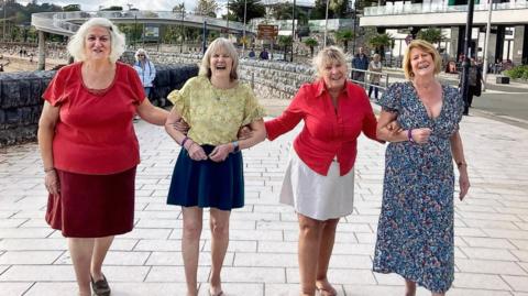 Four women link arms on a seaside promenade and smile at the camera. In the background are steps leading down to a beach. 