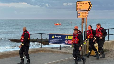 Five men wearing black suits and red lifejackets are walking along the seafront. Some are looking out to sea, some are looking ahead. The sea is visible behind them. An orange search and rescue boat is visible in the distance.