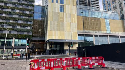 A large hotel building with a golden exterior. There are cones blocking a road below.