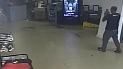 CCTV image of a man wearing a black t shirt and trousers walking towards a machine with a lit up screen inside a building. Racks and shelves can be seen on the left.