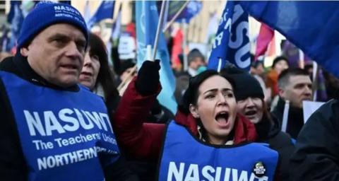 A man and woman are taking part in industrial action. They are wearing blue bibs with 'NASUWT' printed on the front. There are surrounded by other people, some are waving coloured flags. 