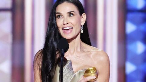 Actress Demi Moore clutches her Golden Globe award to her chest as she makes her thank you speech. She has long dark hair which extends as far as her elbows. and wears a gold dress with bare shoulders. One ear is exposed, from which hangs an ornate earring.