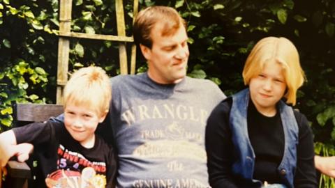 A young boy, older man, and young girl sit on a bench. The older man has his arms outstretched around the children and wears a blue Wrangler t-shirt. The boy has blonde hair and a black graphic t-shirt on, the girl has short blonde hair, a long sleeved black t-shirt and a denim waistcoat over the top. The background of the image features green foliage and a wooden trellis.