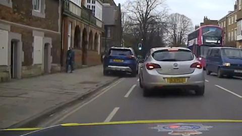 A car is halfway on the pavement as it drives along past queuing traffic