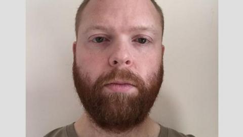 A police mugshot of Paul Chowles wearing a brown T-shirt and standing against a white background. He has a full beard and short red-brown hair, and is looking at the camera with a blank expression. 