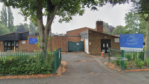 The entrance to a school with gates at the front, two blue signs and hedges, with a one-storey brick building in the background