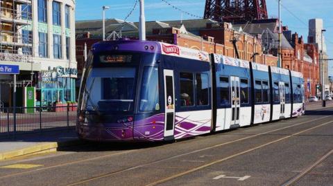 A Blackpool tram, with purple and grey livery, travels along the promenade in front of arcades and the Blackpool Tower