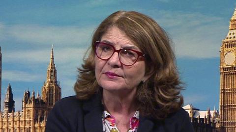 A head shot of a woman in front of an on-screen backdrop of London, showing Big Ben and a blue sky. She has shoulder-length brown hair and is wearing pink-framed glasses, a navy blazer and a patterned blouse.
