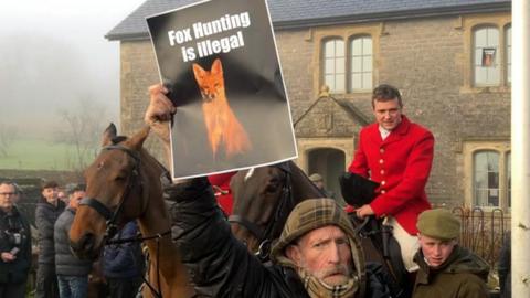 In the foreground is a man wearing a green hooded jumper and black coat holding up a piece of paper with a fox on it and the words 'fox hunting is illegal on it. Behind him is a man on a horse wearing traditional red hunting gear. There is a man in a green flatcap and coat stood next to him. In the background is a brick house and large fields to the left. 