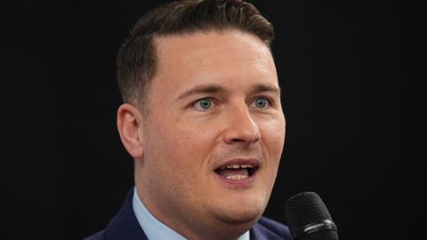 Wes Streeting speaking at an event. He is wearing a blue shirt and jacket and holding a microphone. He has dark hair.