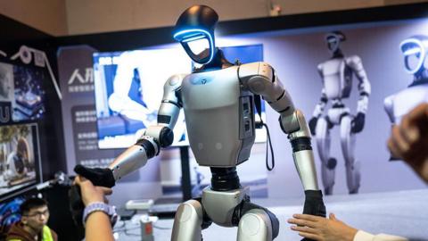 A humanoid robot being demonstrated at an event. With the stance of a human, its arms, torso and pelvis are silver brushed metal, with black joints and black hands. The face is a hollow ring that glows icy blue around the perimeter. In the background is a display board with large images of the same robot, while human hands reach out to shake the robot's black hands. 