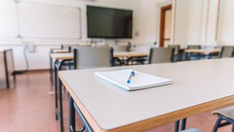 A generic picture of a classroom, with a notepad and pen on a desk in the forefront of the picture.