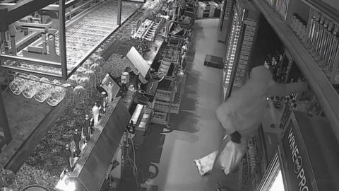 A black and white CCTV image of a man stealing bottles of alcohol from behind a bar in a pub