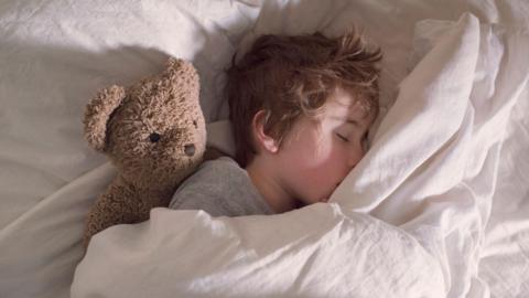 A boy sleeping in a bed which has white sheets and a white duvet. Next to him is a brown furry teddy bear.
