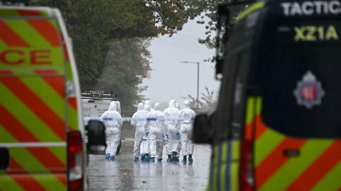 Picture from the scene of the attack. A group of forensic officers in Hazmat suits stand in the street in the middle distance. In the foreground two police trucks are visible. It looks like it has been raining