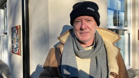 A man wearing a thick jumper, coat, scarf and woolly hat standing outside. He is looking into the camera. He is standing in front of a white wall.