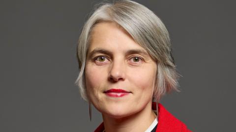 Anna Dixon: a woman with short grey hair wearing a red coat and red lipstick