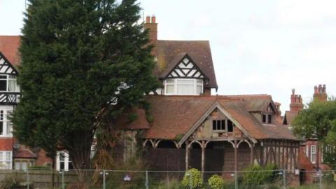A ramshackle Victorian building, flanked by trees and surrounded by a metal fence. Some Tudor-style houses loom in the background.
