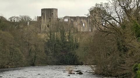 The ruin of a large castle on a hill surrounded trees in front and a river running along the fornt.