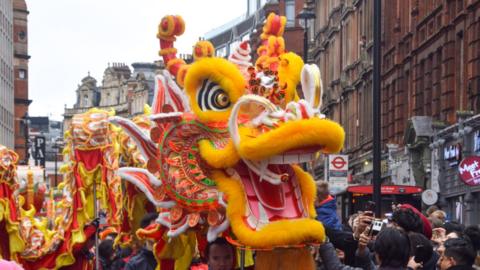 Vibrant dragon costume leading a parade, adorned with bright colours and intricate patterns, surrounded by a cheering crowd.
