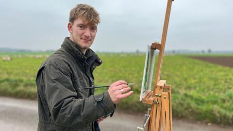 Kieron Williamson smiling at the camera side on, while painting outside. He has short blonde hair and stubble and is wearing a dark green waxed jacket. He is painting on to paper held by an easel. In the background are sheep in a field. It is a dull winter's day. 