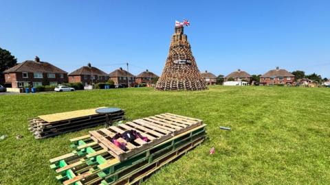 Wide shot of bonfire, built using wooden pallets, at Churchill Park. There are pallets on the grass in the foreground. In the background, there are seven semi-detached, red brick properties.