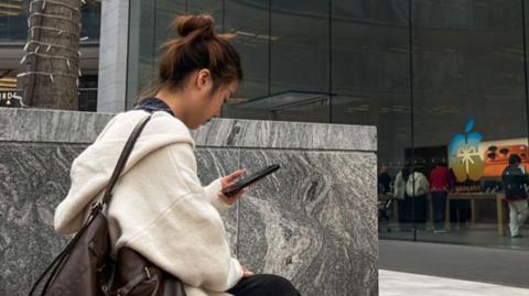 A woman uses a smartphone while sitting outside an Apple Store in Chongqing, China.
