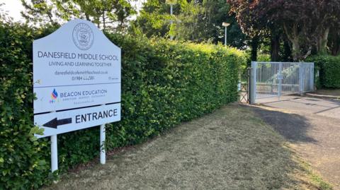 An exterior view of the school showing a hedge and the school sign pointing to the entrance off to the left