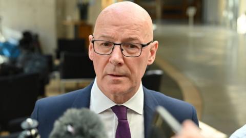 John Swinney, who is bald with glasses, speaks into microphones. He is wearing a business suit 