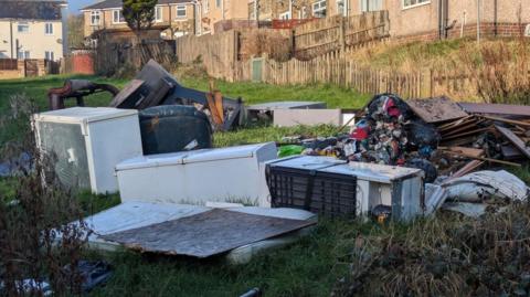 A fly-tip on a patch of grass, which borders some houses in the background. The fly-tip comprises white goods, furniture, planks of wood and other rubbish.