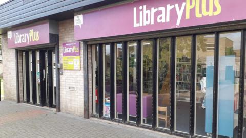 The outside of the library, which has large floor to ceiling glass windows and a purple sign saying Library Plus