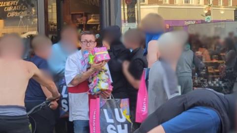 A man wearing an England flag t-shirt carries an armful of Lush gift sets looted from the shop. There are more people heading into the store, whose faces are blurred out.