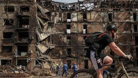 A cyclist rides past a damaged building, with a crew cleaning up in front of it