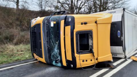 An overturned lorry with a yellow cab, the windscreen is smashed