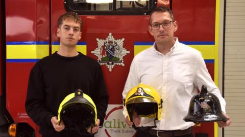 Ben and Steve Wright stand in front of a fire engine. They are holding three helmets - one is Ben's, one is Steve's and one belonged to Steve's father Nick