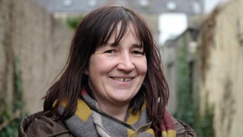 Aisling is smiling at the camera and has shoulder length brown hair with a fringe. She is wearing a grey and yellow scarf.