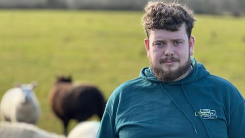 A man standing looking at the camera with a neutral expression. He is wearing a blue hoodie. He is standing in a field with some sheep behind him.