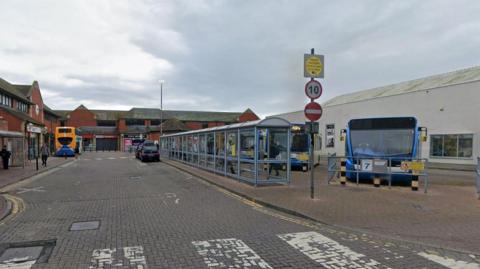 A Google Streetview of the bus station in Carlisle. It is a wide cobbled dead-end street with different bays with bus shelters on one side and parking bays for buses on the other side. There are some shops at the bottom of the road.