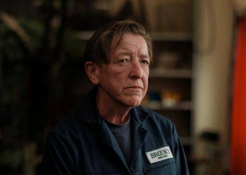 Lisa looks ahead, side-on to the camera. She has short dark hair and is wearing a blue top and blue overalls, bearing the name Brooks.