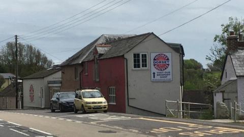 A street view of the former Dorset knob factory. It is a row of four properties in varying heights, painted red, cream and brown on the front and with a grey tile roof. On the side is a sign for the factory which has a circular logo for the biscuits on it, it reads 'Moores Dorset Biscuits. Visit out Bridport shop'. There are cars parked out the front of the building and a section of the road can be seen to the left of the image.