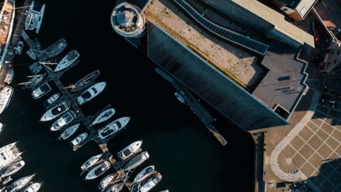 A drone shot of a building next to a small harbour, with boats lined up next to jetties.